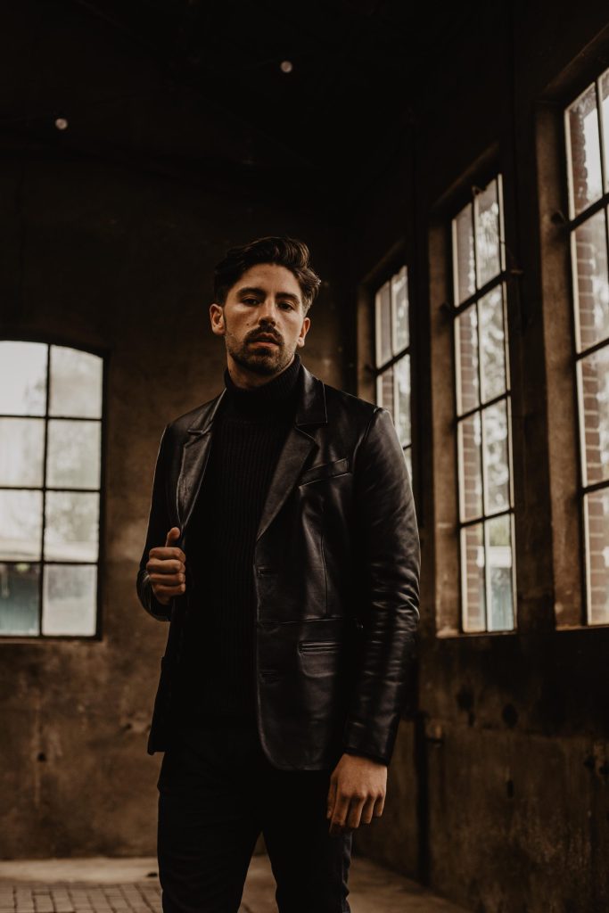 Man in a dimly lit industrial setting wearing a black leather blazer over a turtleneck sweater, gazing forward.